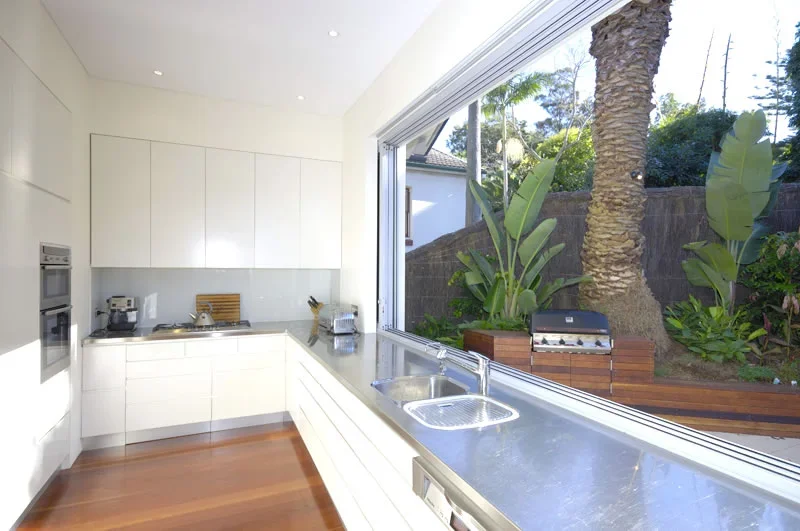 Modern kitchen with white cabinets, stainless steel appliances, and a large window overlooking a garden with tropical plants and a tree.