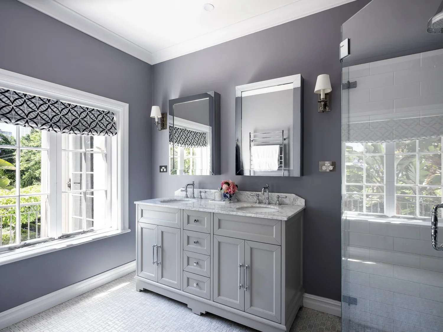 Bathroom with gray walls, white cabinetry, and twin sinks with marble countertops. Two mirrors above each sink, with wall-mounted lights on either side. A window with a patterned black and white Roman shade letting in natural light, and a glass-enclo