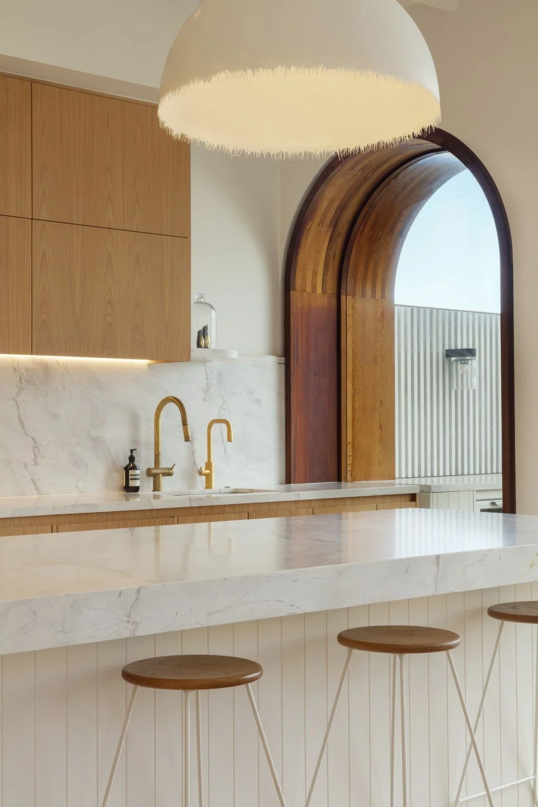 Modern kitchen with white marble countertop, gold fixtures, wooden cabinets, and a large arched window.