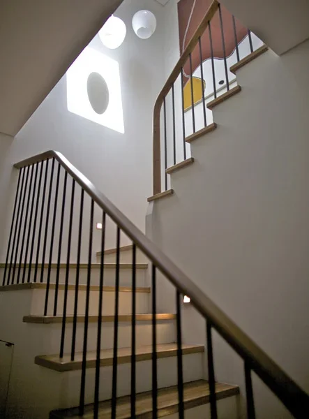 Interior view of a staircase with wooden steps and black metal railings, illuminated by natural light through circular and square windows on the white wall.