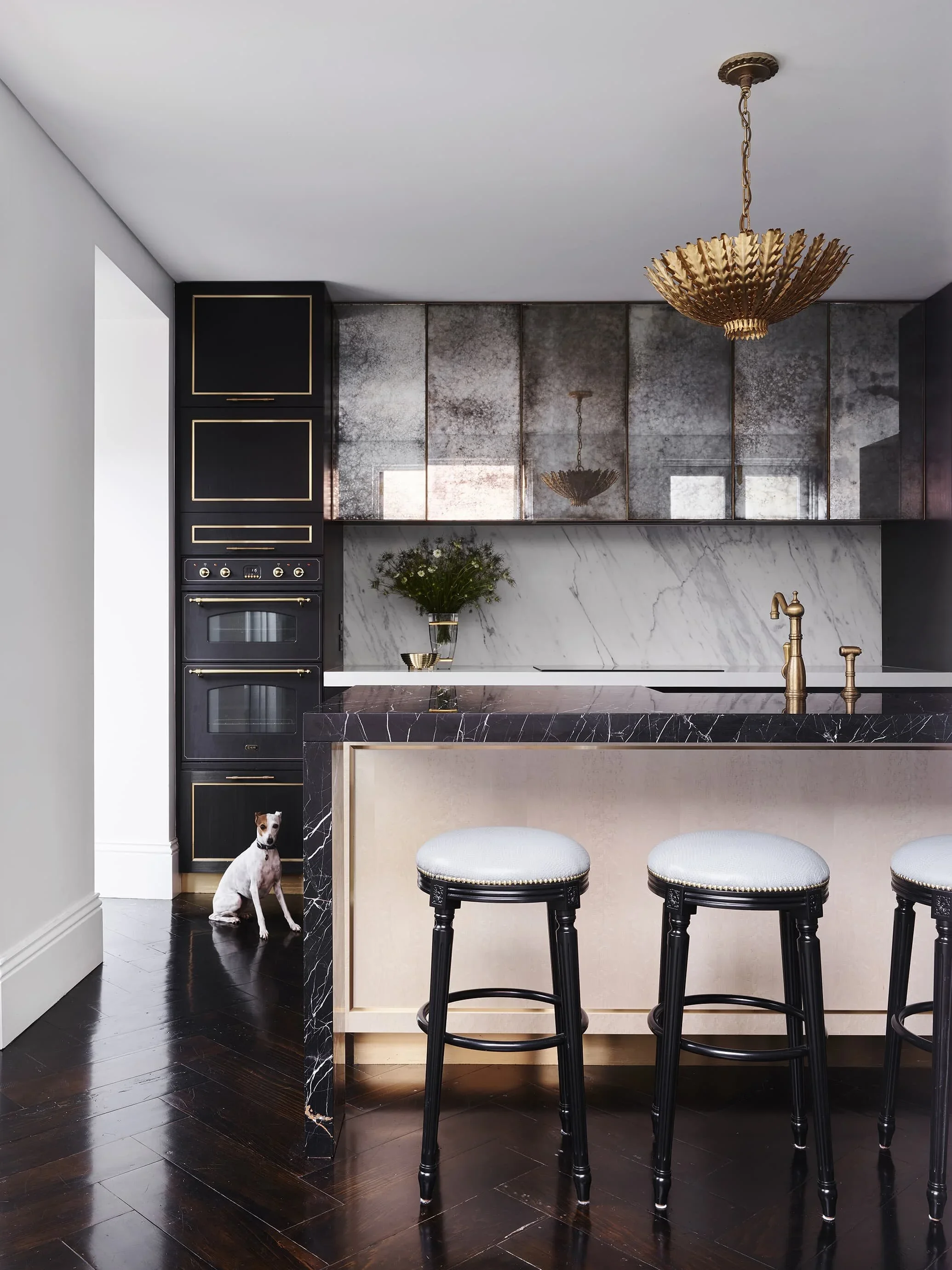 Modern kitchen with black, gold, and marble accents. Features a marble island, brass fixtures, black and gold cabinetry, and three black barstools with white seats. A small dog sits on the dark hardwood floor near the corner.