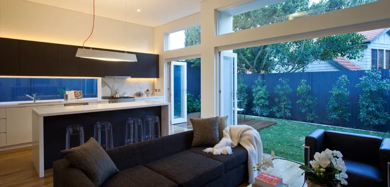 Modern open-concept living room and kitchen with large windows, a lounge sofa, and a view of a backyard with trees and a fence.