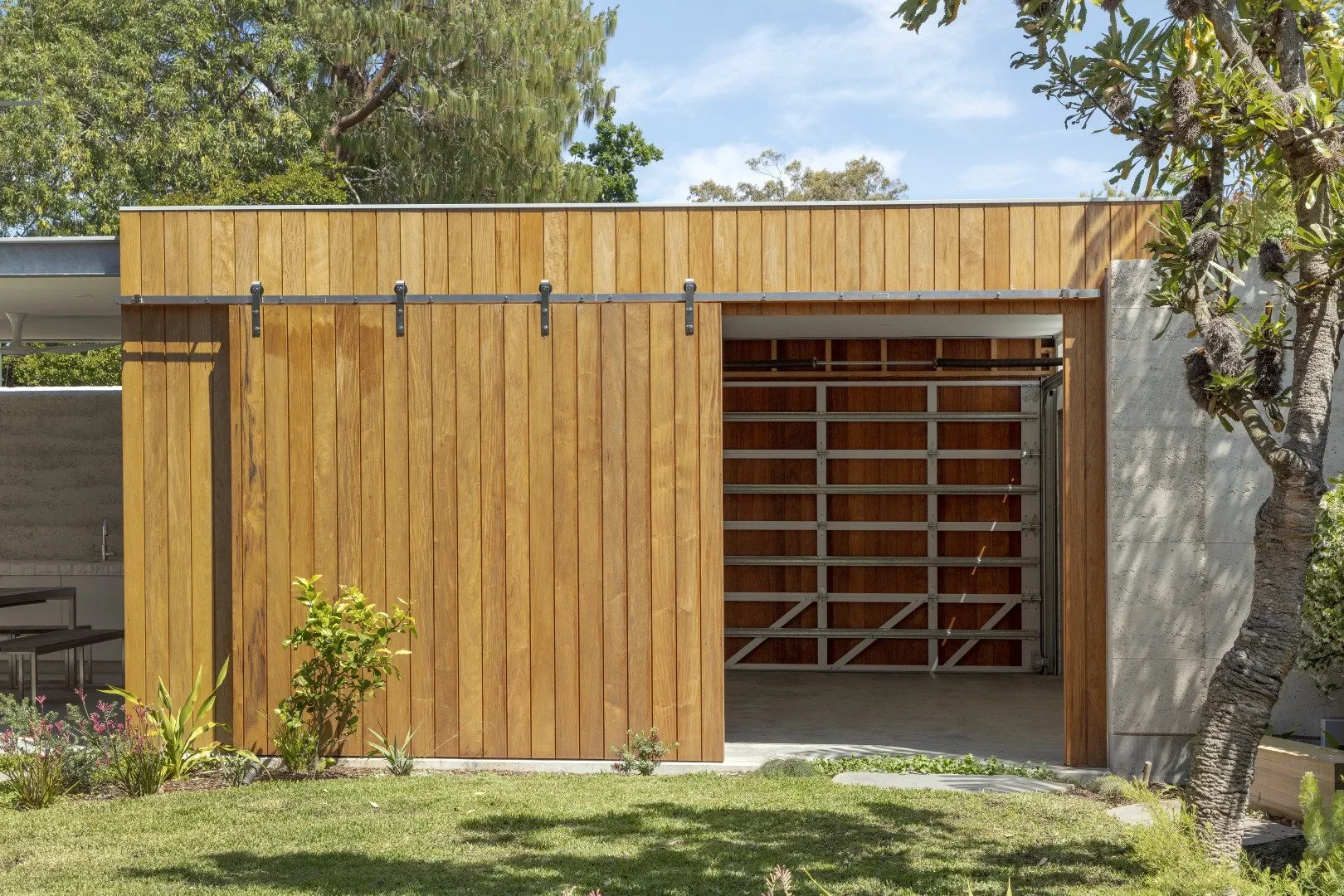 A modern garage with a wooden sliding door, an open section showing a metal garage door, surrounded by trees and a well-maintained lawn.