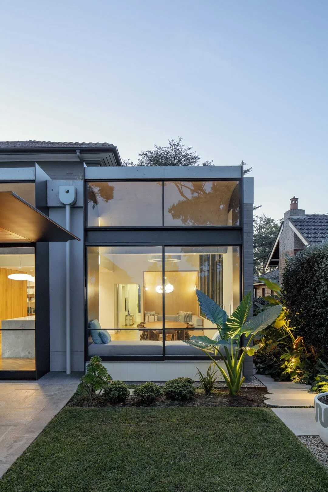 Modern house with large glass windows, a small front garden, and lush tropical plants, during dusk with interior lights on.
