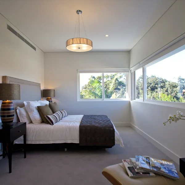 A bedroom with a large window showing green trees outside, a bed with white bedding and striped pillows, dark nightstands with matching lamps, a small table with magazines, and a ceiling light fixture.