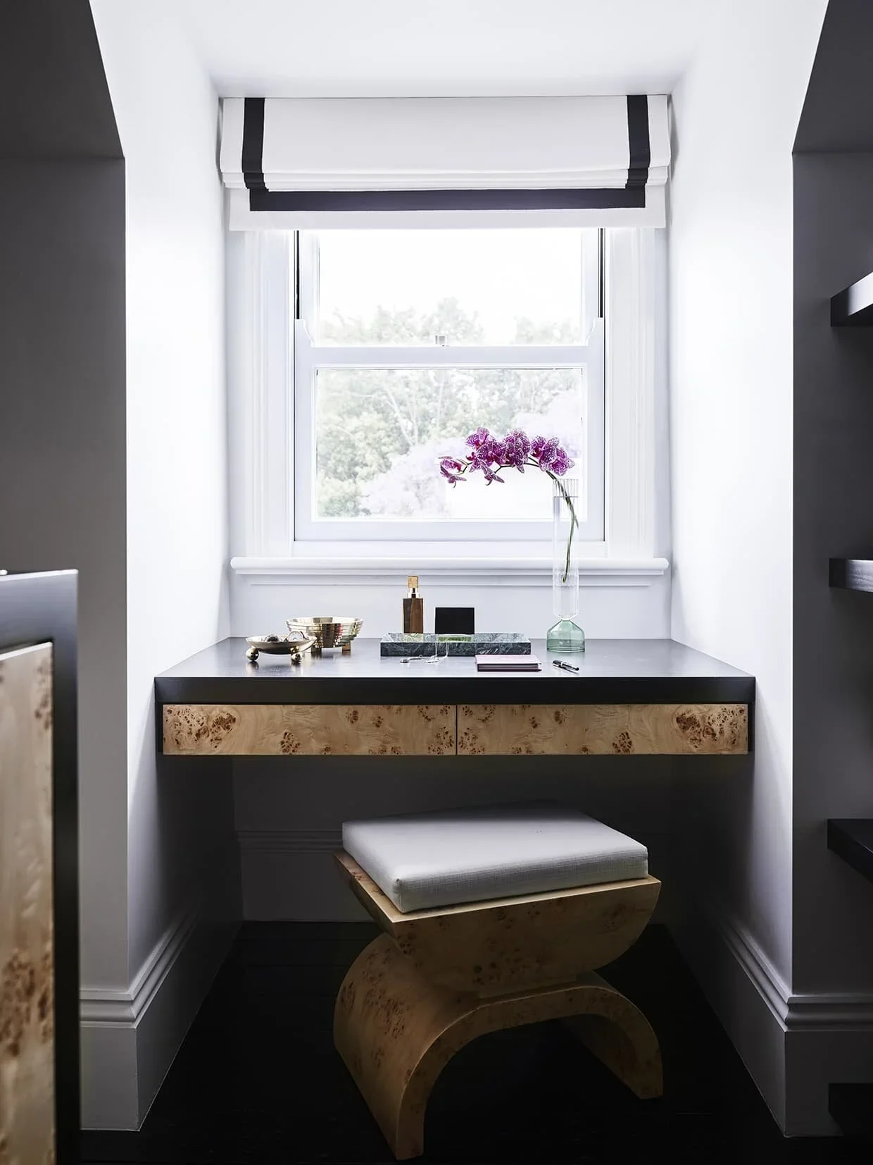 A small desk with a black surface and light wood drawers beneath a window. On the desk are a perfume bottle, a tray with small items, and a vase with purple flowers. There is a matching light wood and padded stool in front of the desk.