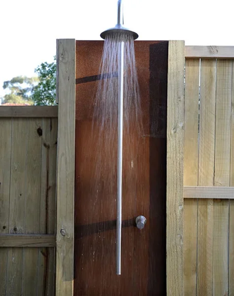 An outdoor shower with water running from a metal showerhead mounted on a vertical pipe, installed on a wooden privacy wall.