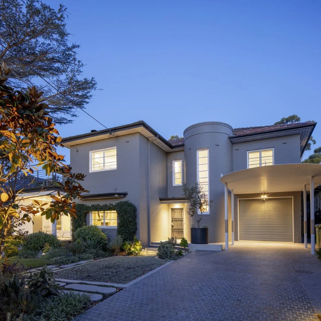 Modern two-story house with a landscaped front yard and driveway at dusk, illuminated from inside.