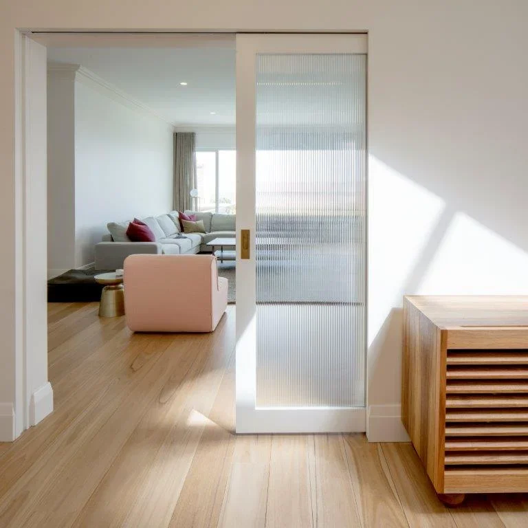 View of a modern living room seen through a sliding glass door, with a white sofa, pink armchair, and wooden furniture, bright natural light, and hardwood floors.