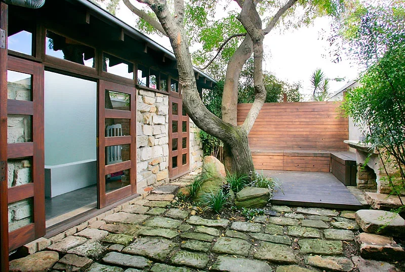 A backyard patio with a cobblestone ground, a large tree, and a wooden privacy wall. There is a built-in bench and a stone planter with plants.