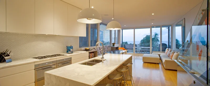 Modern open-plan kitchen and living room with white cabinetry, marble island, pendant lights, hardwood floors, and large windows with a view.