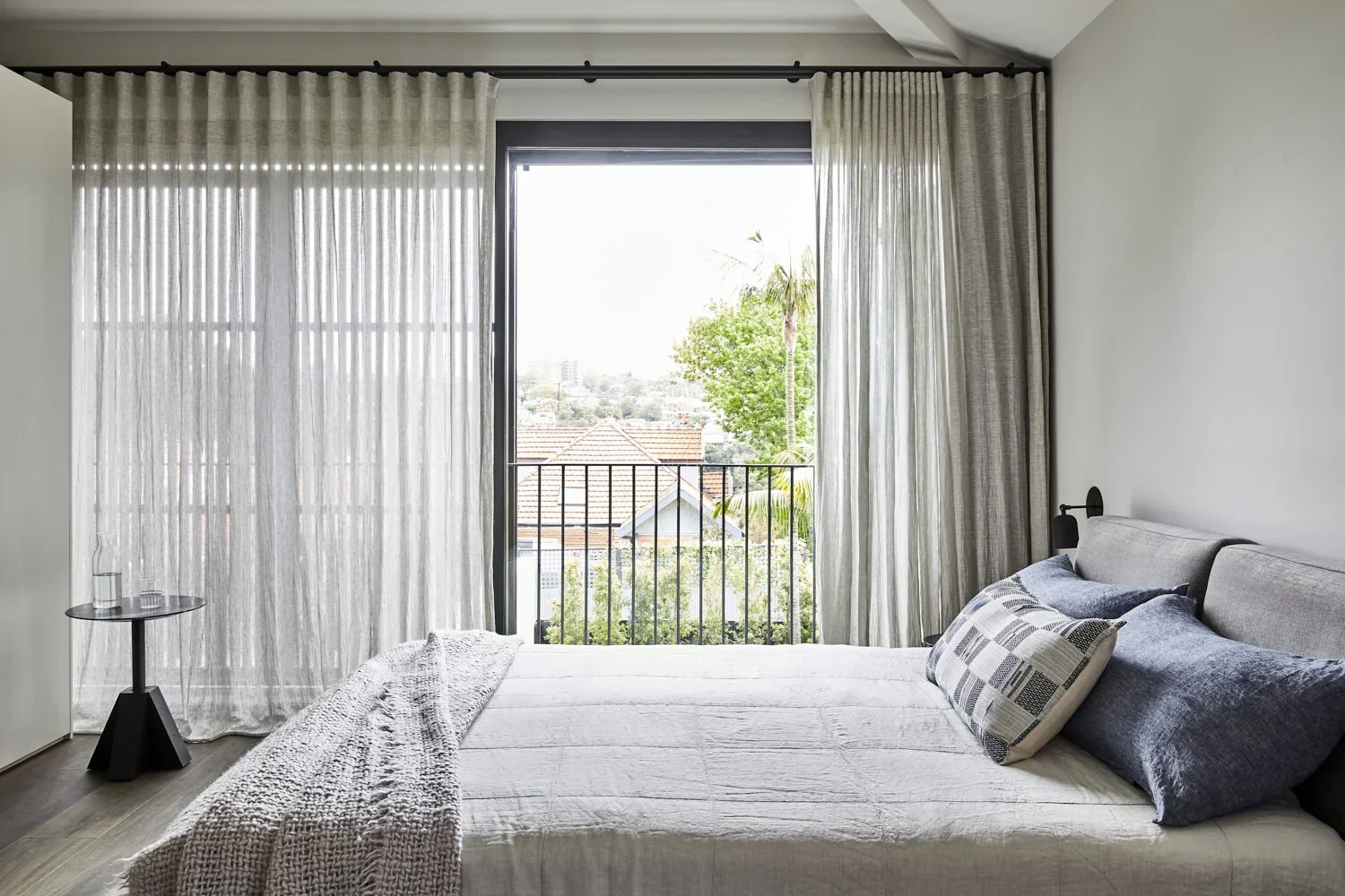 Bedroom with large window, sheer curtains, bed with white bedding, and pillows, balcony with view of trees and rooftops.