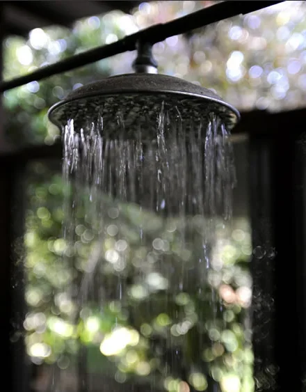 A close-up of an outdoor showerhead with water flowing from it, against a background of blurred trees and sunlight.