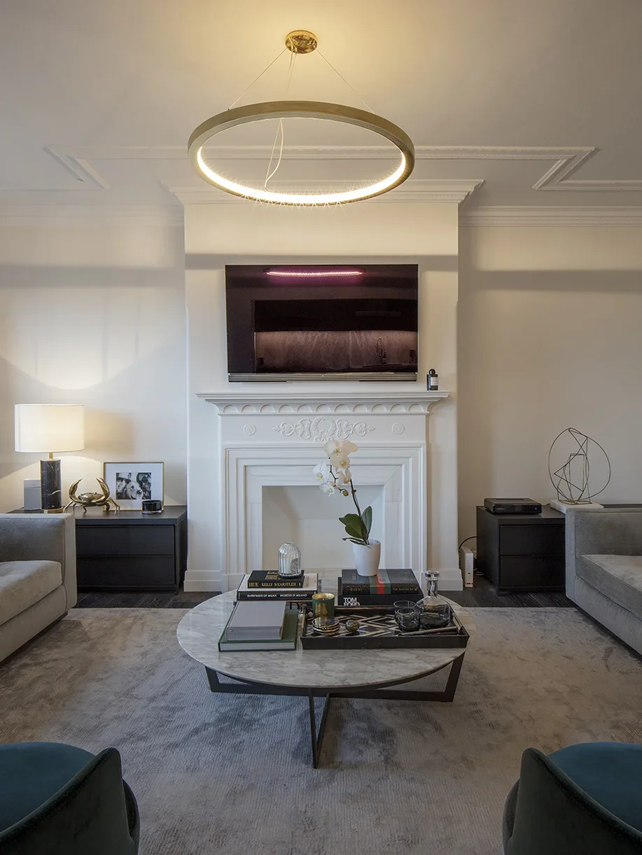 Living room with a modern chandelier, white fireplace, wall-mounted TV, two gray sofas, a coffee table with books, a potted orchid, and decorative objects.