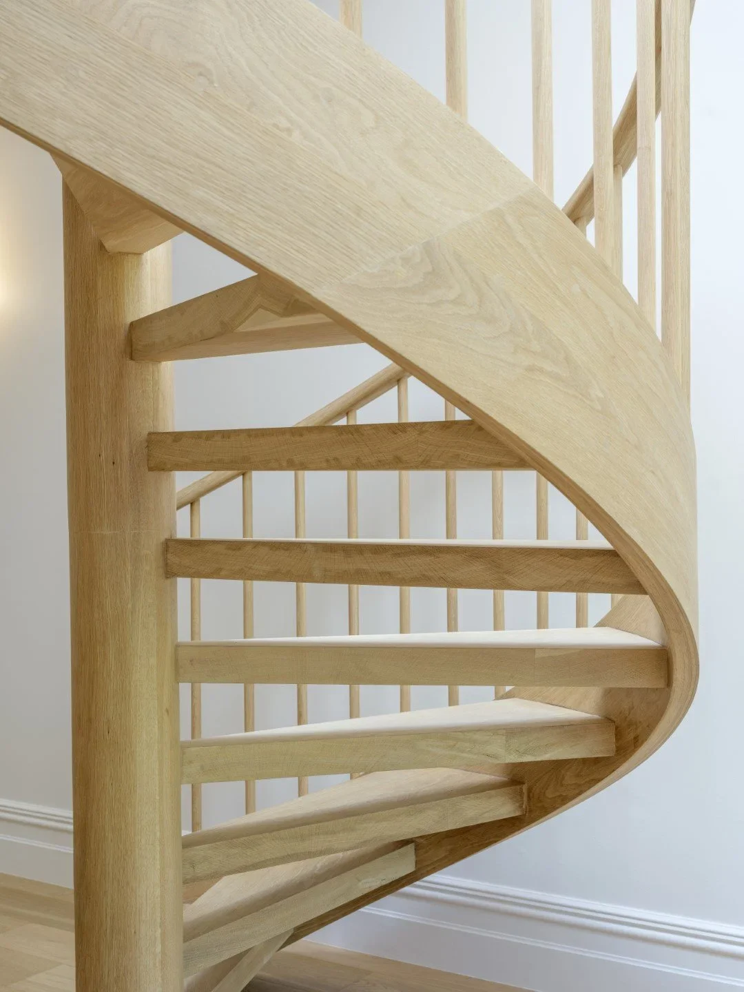 Close-up of a wooden staircase with a curved handrail and vertical balusters, photographed against a light gray wall.