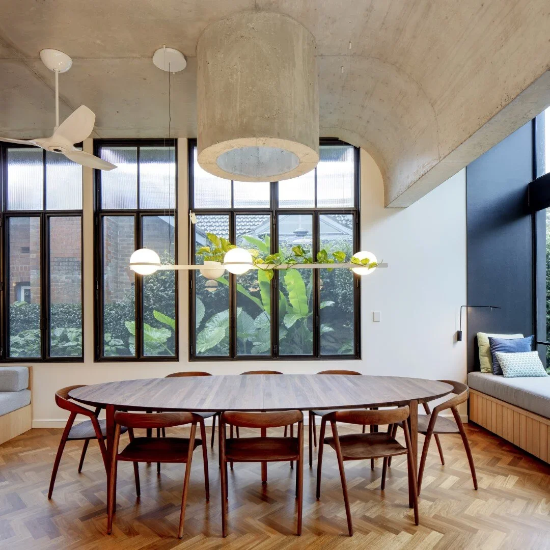 Modern dining room with a wooden oval table surrounded by nine wooden chairs, large windows with black framing, green plants outside, and contemporary lighting fixtures hanging from a concrete ceiling.