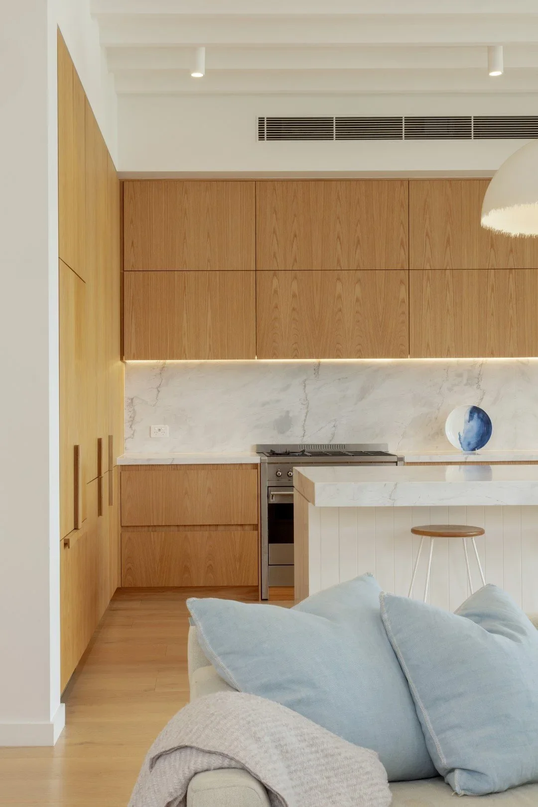 Modern kitchen with wooden cabinets, marble backsplash, and a bar counter with a stool. A decorative blue and white plate is on the counter. Part of a light-colored sofa with blue pillows and a beige throw is visible in the foreground.