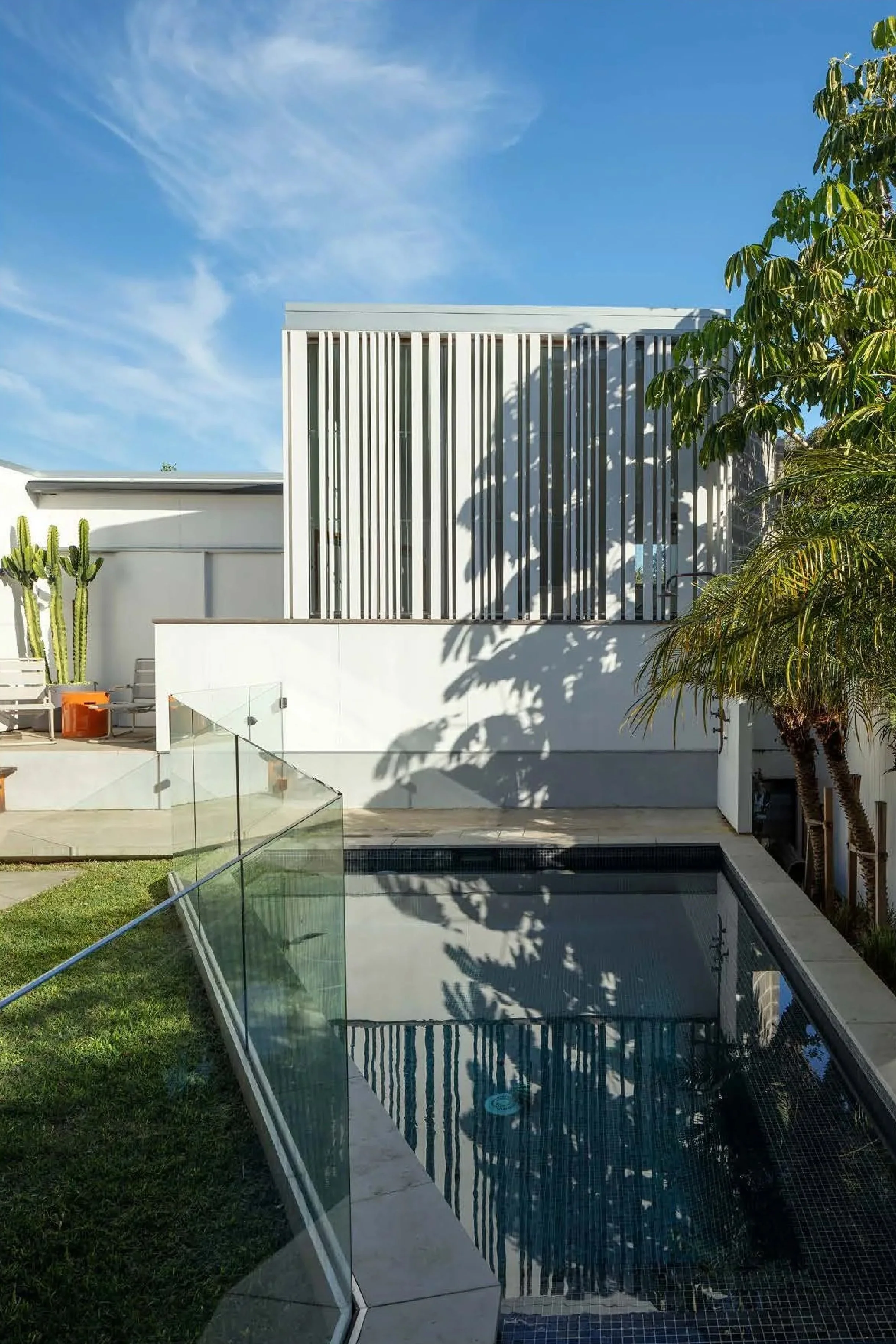 Modern house with white exterior walls, a small pool with a glass fence, and outdoor seating area featuring a cactus plant and trees, under a blue sky with wispy clouds.