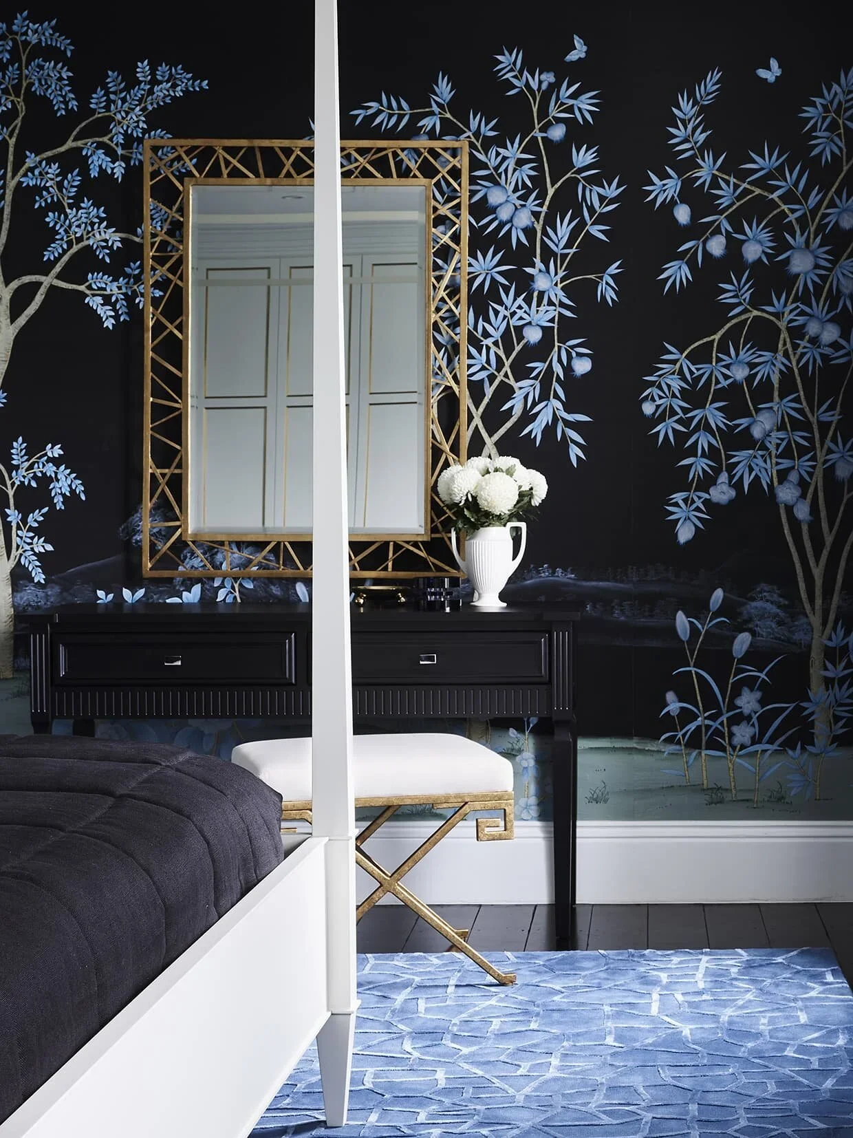 A bedroom corner with a white bed frame and a dark cover, a black dresser with a white flower vase, a gold-framed mirror, and a wall with a blue floral and tree pattern mural.