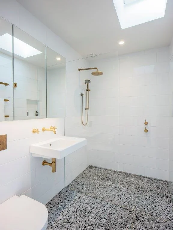 Modern bathroom with white tiled walls, a floating sink with gold fixtures, a mirror, a shower with gold fixtures, and a terrazzo floor. There's a skylight overhead and recessed lighting.