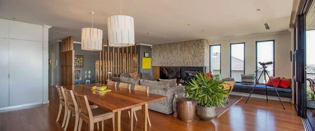 Modern living room with hardwood floors, a large gray sofa, a wooden dining table with six chairs, a large potted plant, a telescope near windows, and colorful cushions on built-in window bench.