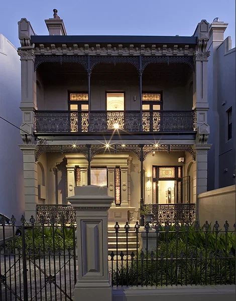 Two-story Victorian-style house with decorative ironwork balconies, front porch, and a small garden enclosed by a black metal fence.