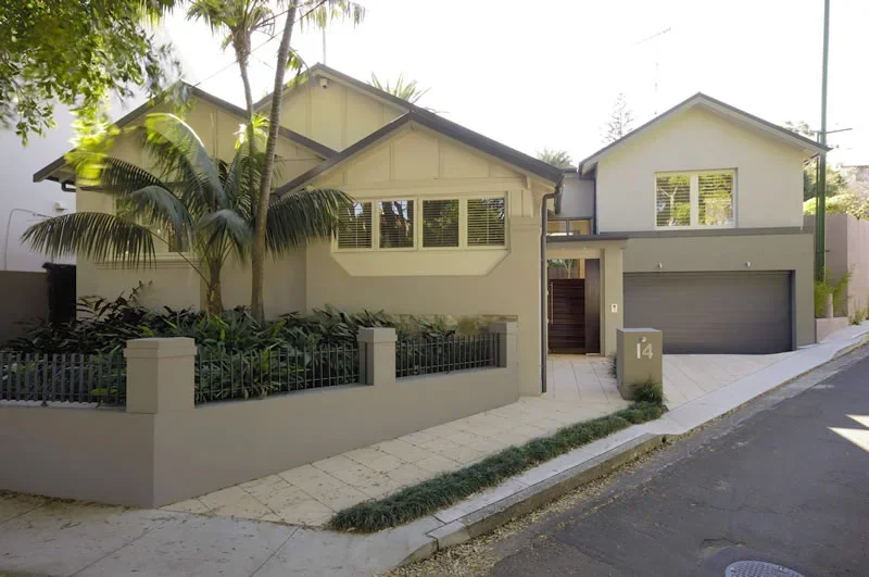 Modern two-story house with a light-colored exterior, front yard with plants and a low fence, large windows, and a gray garage door.