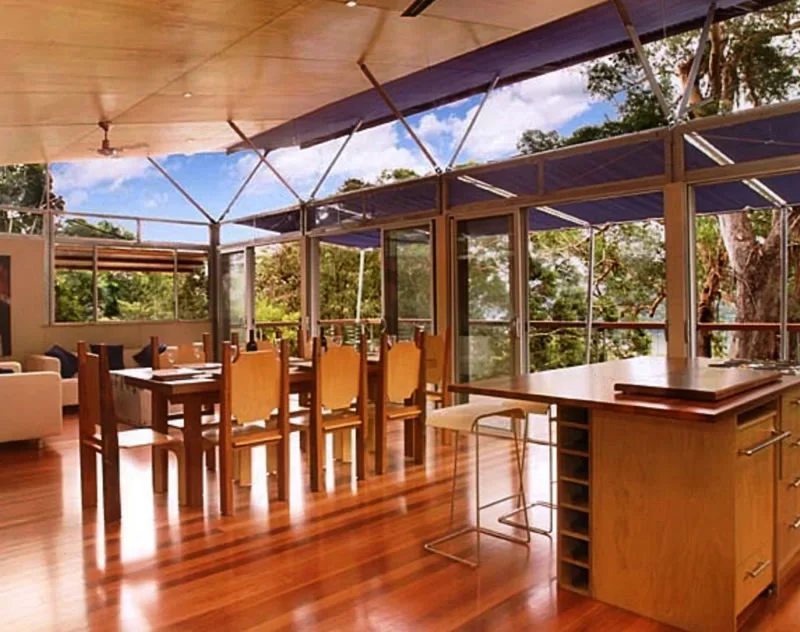 Bright dining room with wooden flooring, glass sliding doors, and a view of trees outside; furnished with dining tables, chairs, and a kitchen counter.