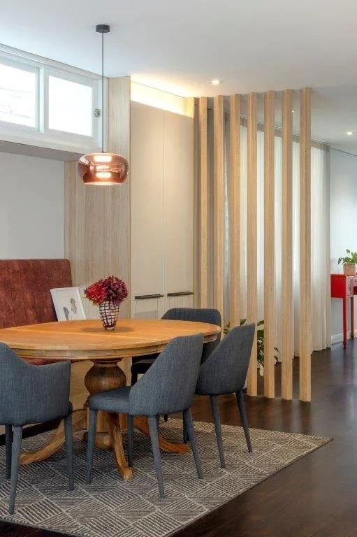 Dining area with a round wooden table, four upholstered chairs, a hanging copper pendant light, a vase with red flowers, and a decorative wooden room divider in a modern interior.