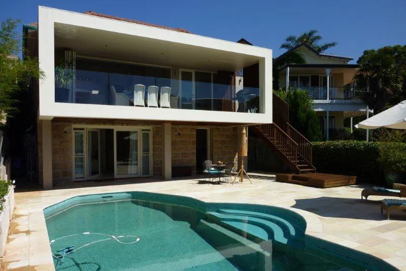 Modern two-story house with a pool in the backyard, glass balcony, outdoor seating, and umbrella, with neighboring homes and lush trees.