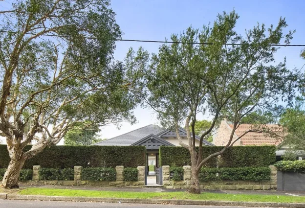 A residential house behind a stone and hedge fence, with two large trees in the foreground and a clear blue sky.