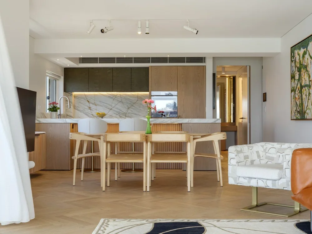 Open-concept living space with dining area, kitchen, and seating. Wooden dining table with chairs, floral centerpiece, marble kitchen backsplash, modern cabinetry, and a colorful abstract painting on the wall.
