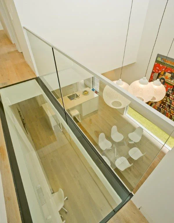 A modern interior view looking down from above, showing a glass railing, a small dining area with four white chairs around a glass table, a kitchen counter with sink and some items, and a wooden floor.