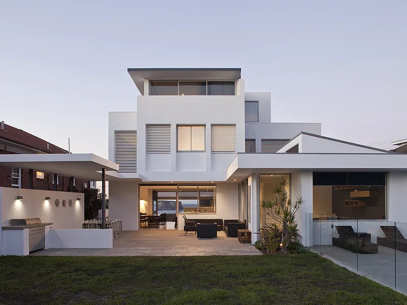Modern white multi-story house with outdoor patio, seating area, and glass railing, during early evening