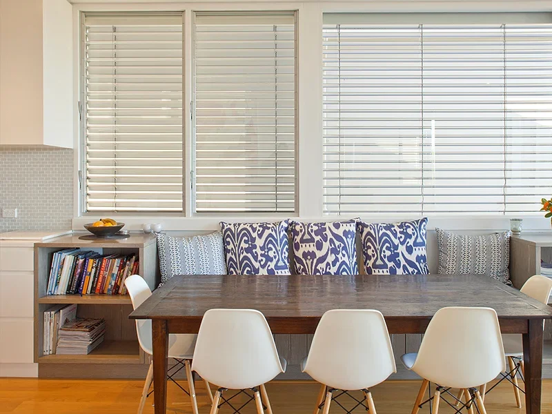 Dining area with a wooden table, six white chairs, a window with blinds, pillows on a built-in bench, and a bookshelf.