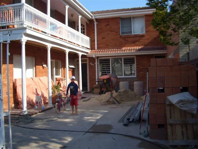 Two children, one boy wearing a helmet, standing in a backyard under construction, with building materials and equipment nearby.