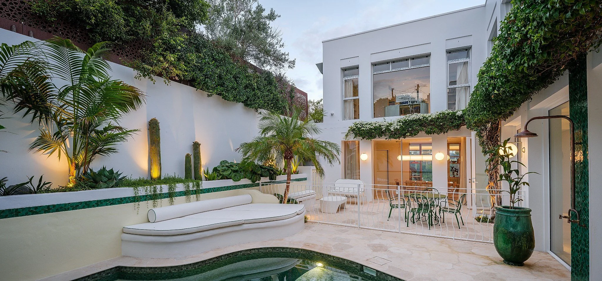 A modern white residential building with large windows, outdoor seating area, a swimming pool, lush green plants, and string lights, during dusk.