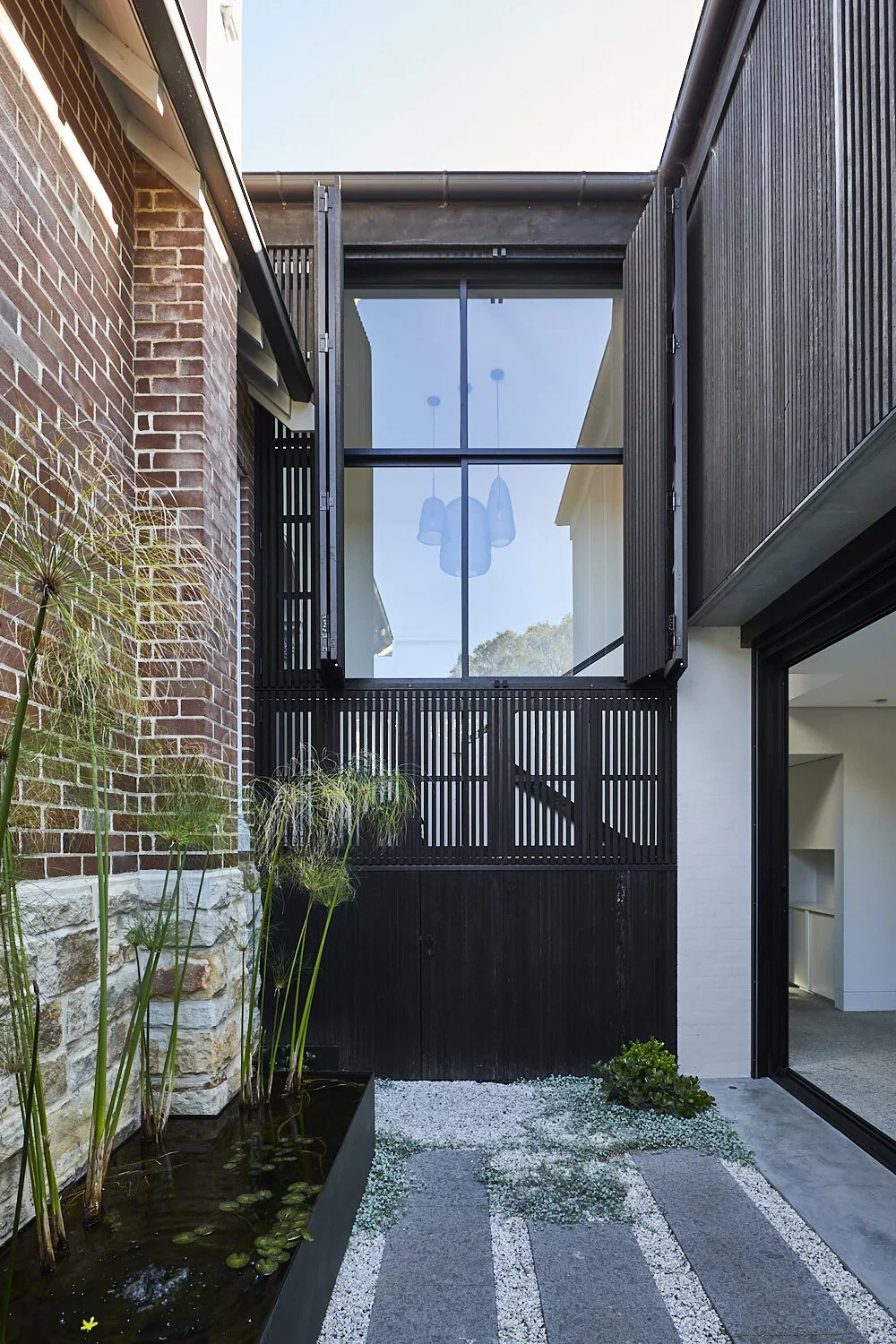 Modern house with a small outdoor patio, featuring a black wooden gate, tall plants in a water feature, and a large glass window with black shutters.