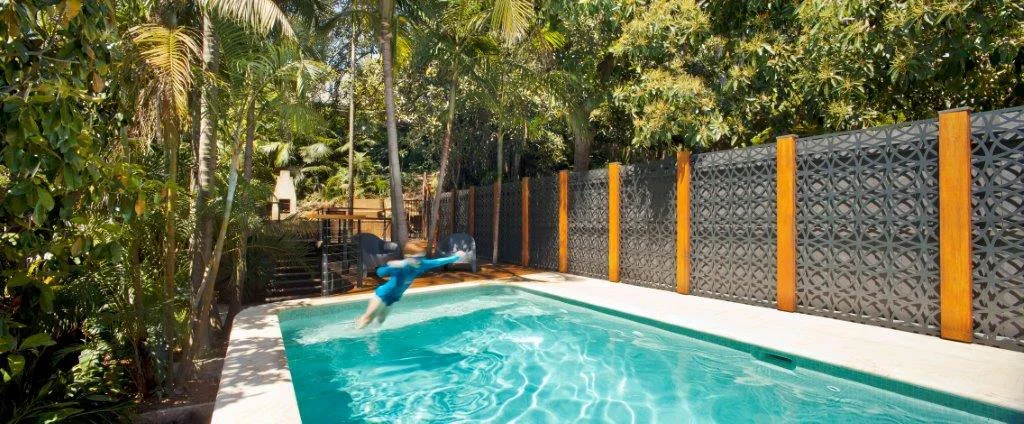 A backyard swimming pool surrounded by lush tropical plants and trees, with a decorative black fence and colorful vertical wooden posts.