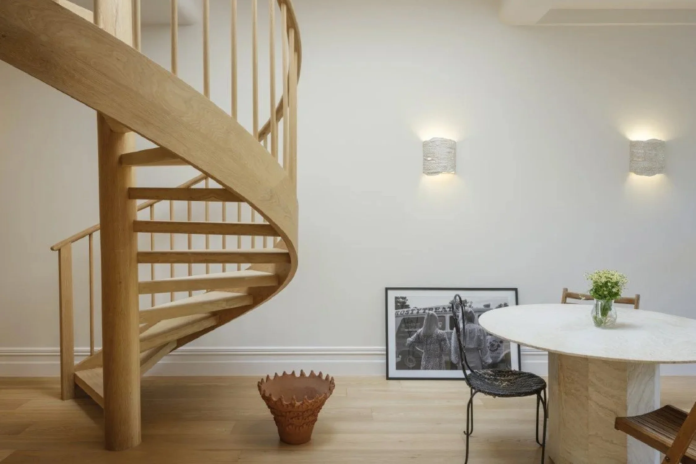 Interior of a room with a wooden spiral staircase on the left, a framed black and white photo on the floor, a round table with a small potted plant, and chairs against a light-colored wall with two wall sconces.