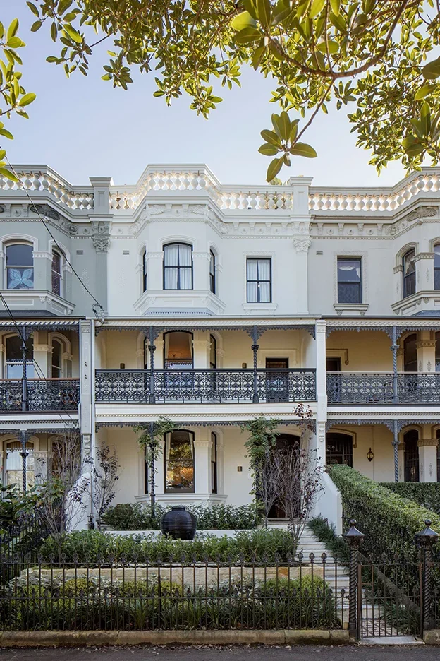 A Victorian-style house painted white with two stories, decorative railings, and large windows, surrounded by a small garden enclosed by a fence.