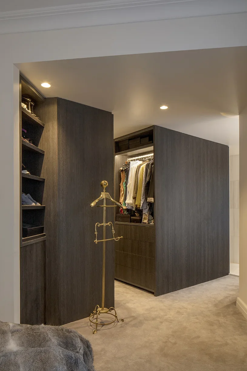View of a walk-in closet with dark wooden cabinets and shelves, clothing hanging on a rod, and a gold coat rack in the foreground, with beige carpeted floor and ceiling lights.