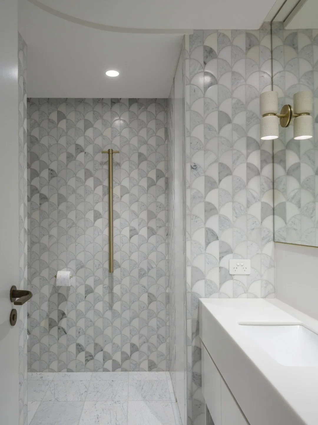 Decorative bathroom with patterned marble tiles, a gold grab bar in the shower area, a white vanity with a sink, and a mirror with wall-mounted cylindrical light fixtures.