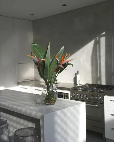 Kitchen with white island and large glass vase with green leaves and orange flowers, sunlight casting shadows.