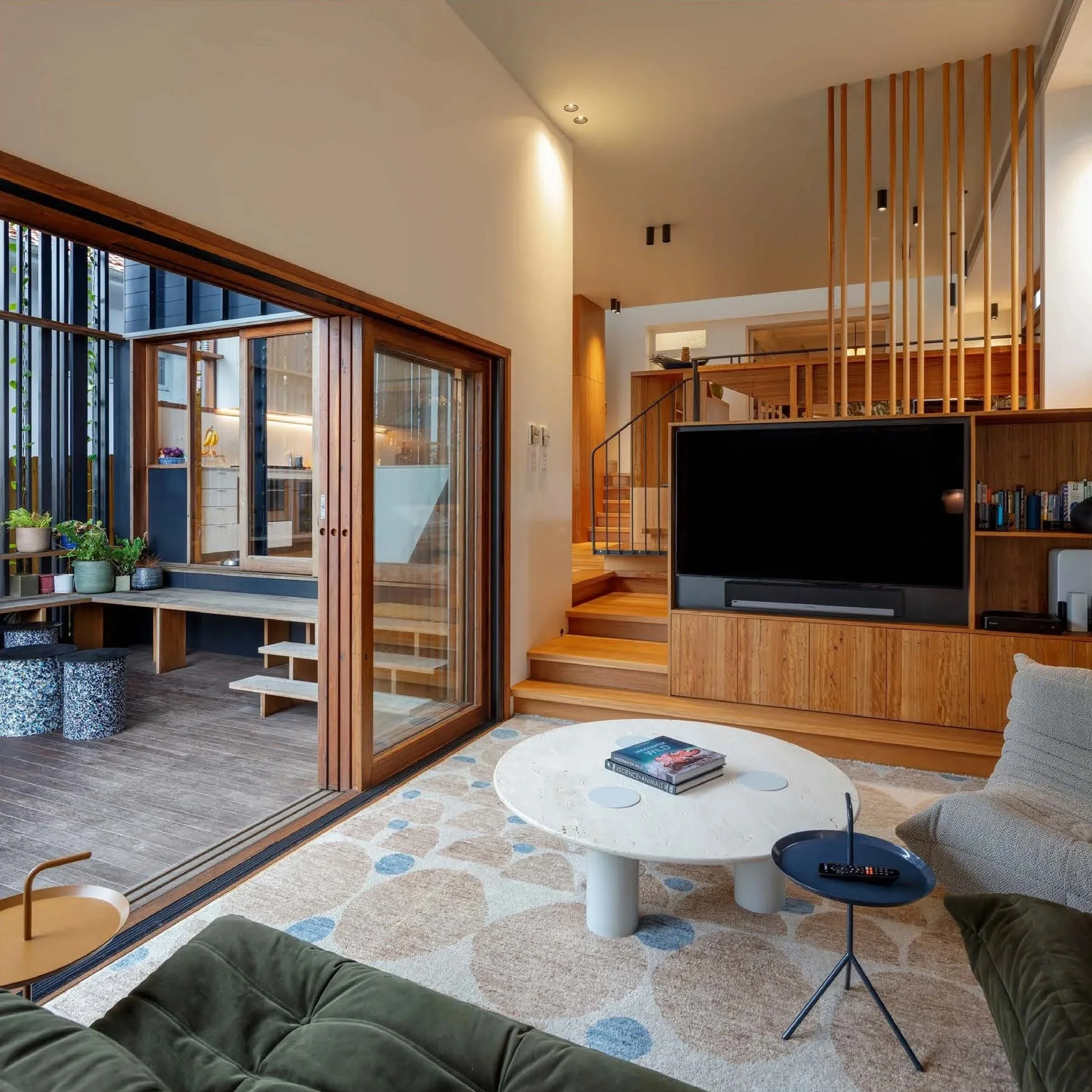 Modern living room with a white round coffee table, a gray sofa, and a black side table with a remote. Large sliding glass doors open to a small outdoor area with a built-in bench and potted plants. Wooden accents and stairs lead to an upper level.