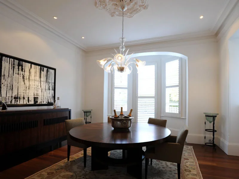 A dining room with a round wooden table, four chairs, a chandelier, a sideboard with artwork, and large windows with shutters.