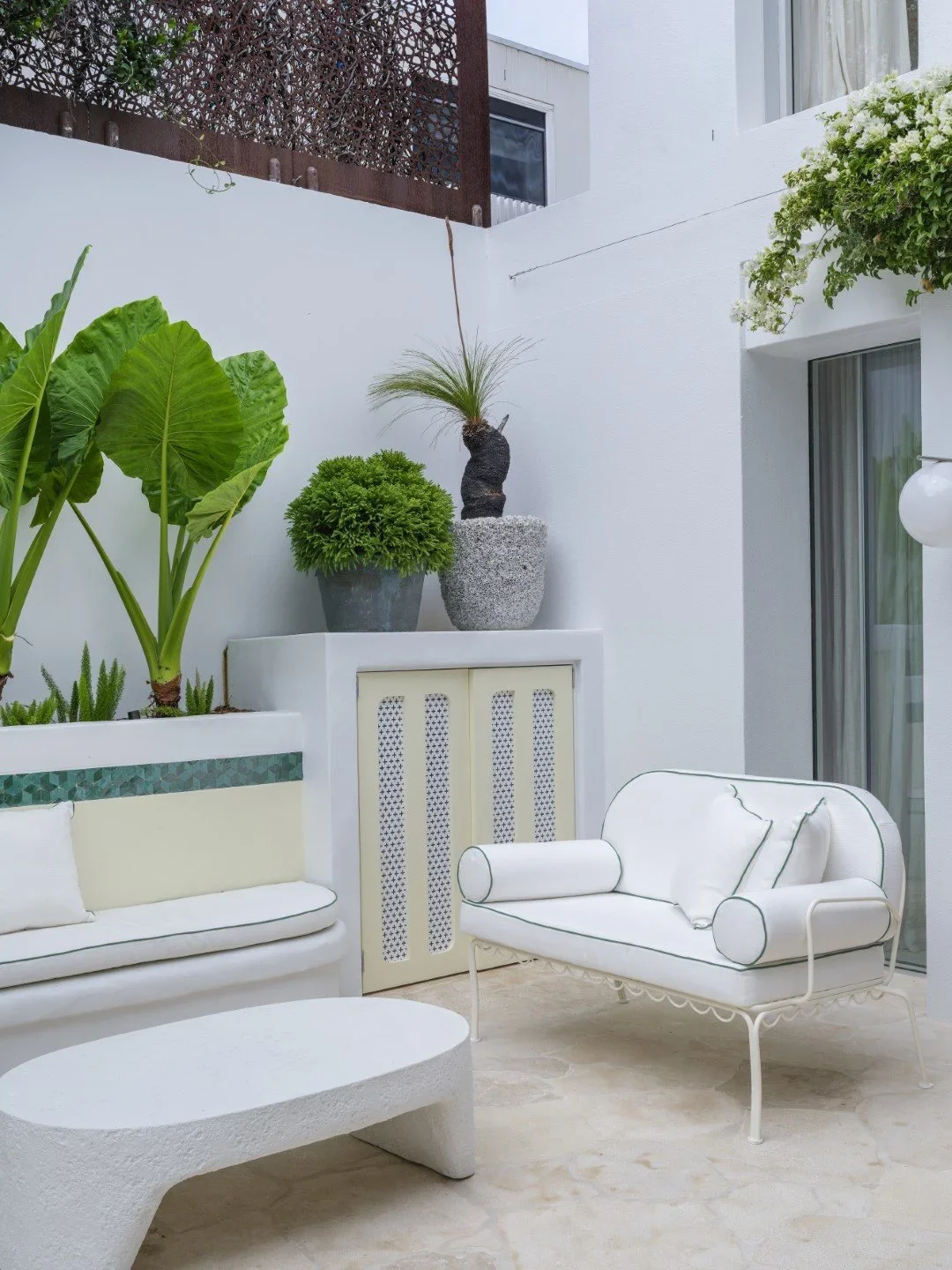 Outdoor patio with white furniture, potted plants including large green leaves, a round white coffee table, and a white wall with a sliding glass door.