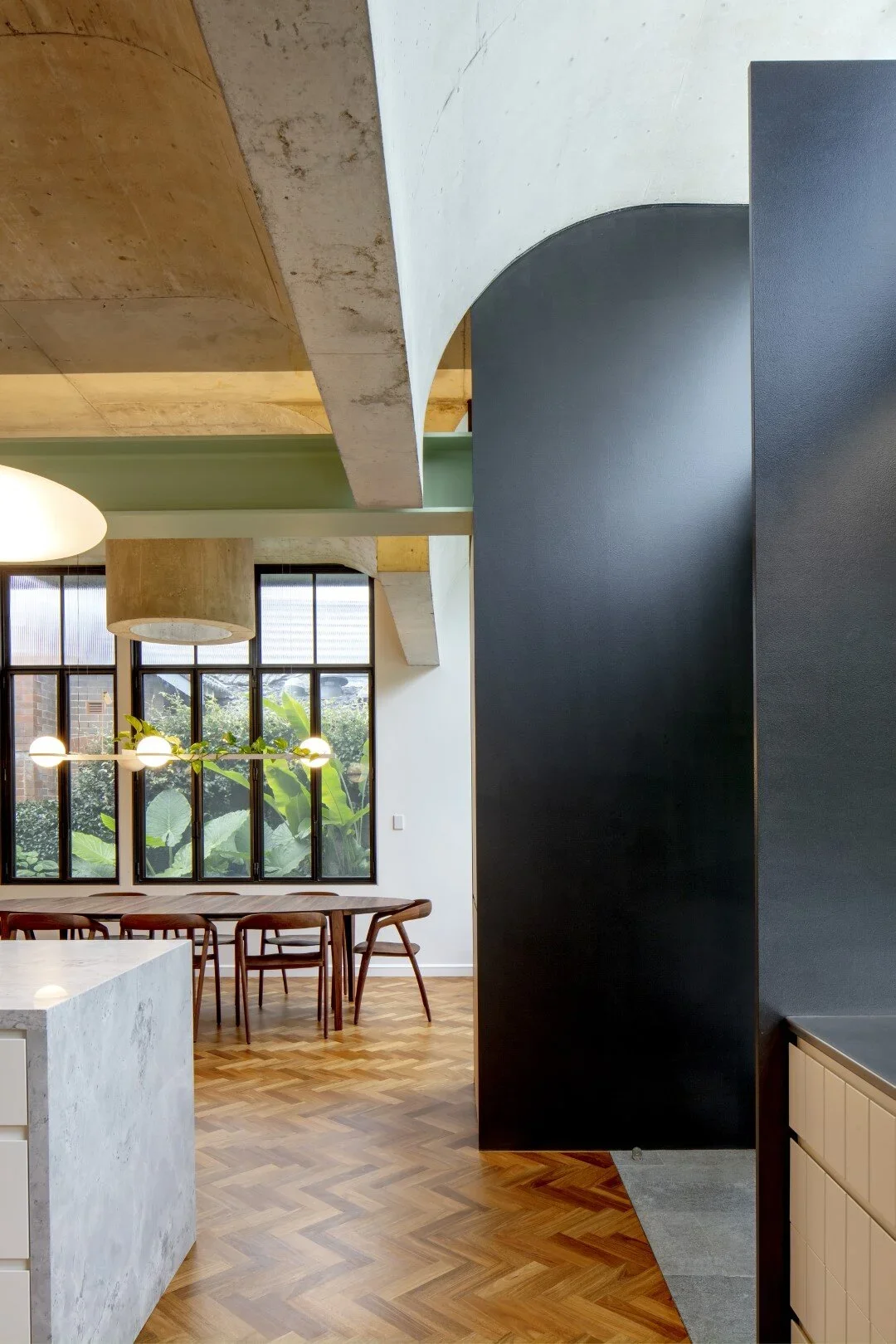 Modern interior space featuring a dining area with a wooden table and chairs, large window with greenery outside, black wall with an arched opening, hanging circular light fixtures, and a mix of concrete, wood, and painted surfaces.