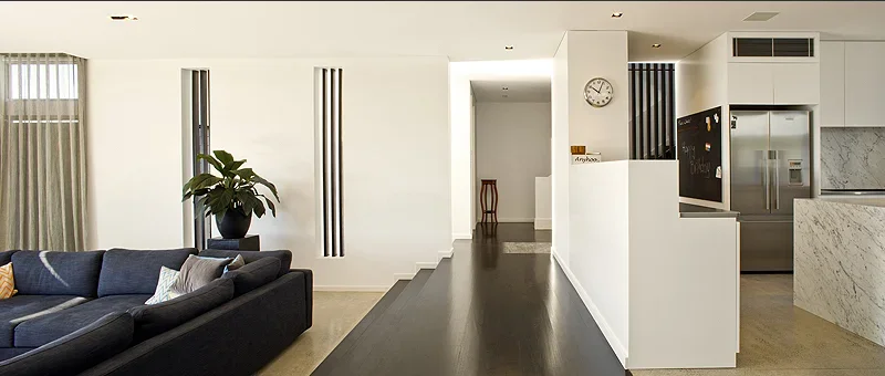 Modern living space with a black sofa, potted plant, wall clock, and kitchen area with stainless steel appliances.