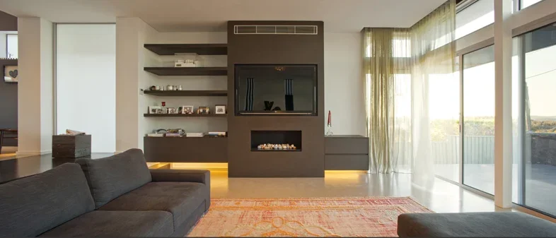 Modern living room with gray sofa, built-in fireplace, wall-mounted television, open shelves, large windows with sheer curtains, and a balcony with outdoor view.